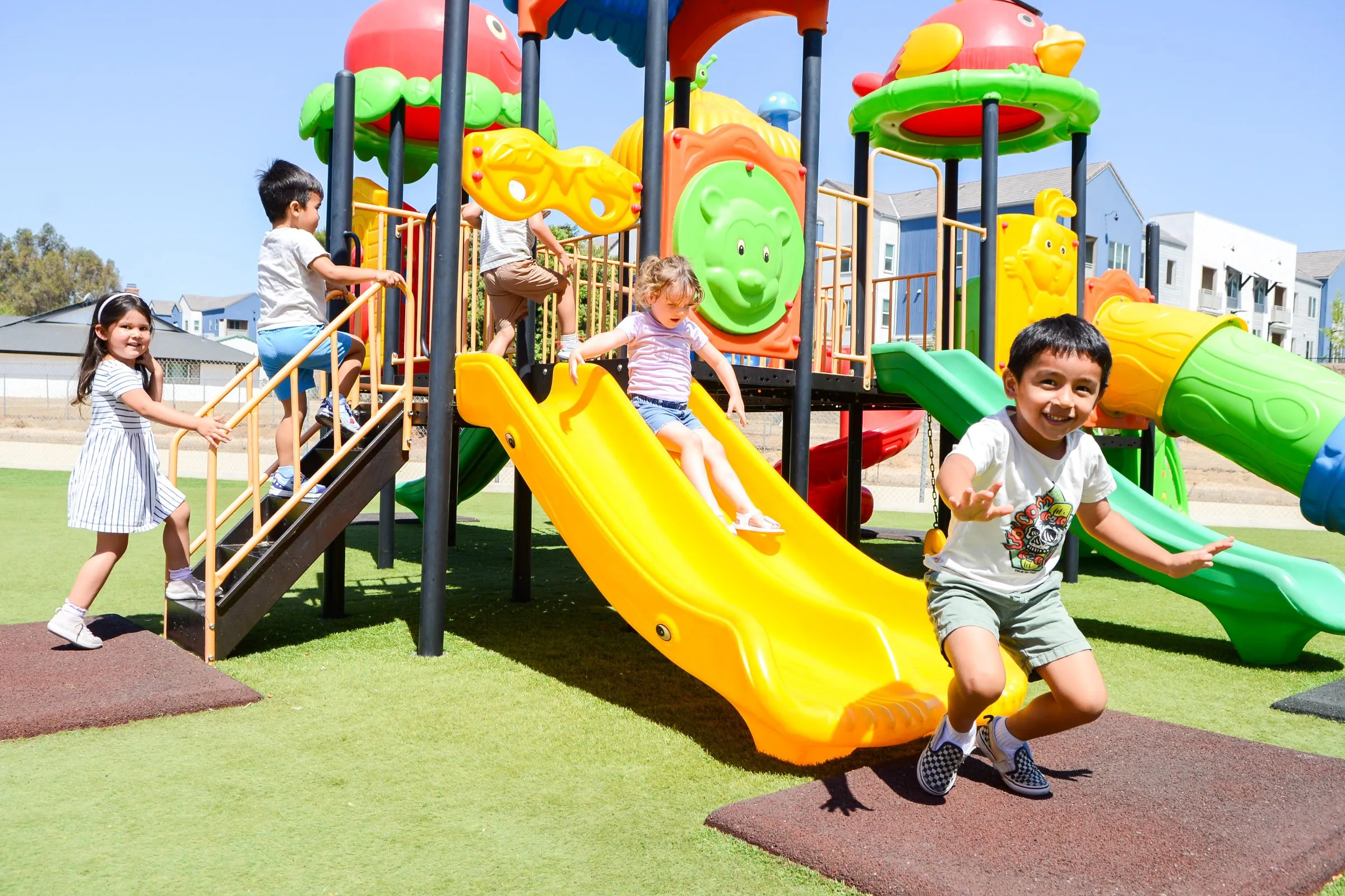 A smiling boy jumps off the end of a yellow slide on the school playground.