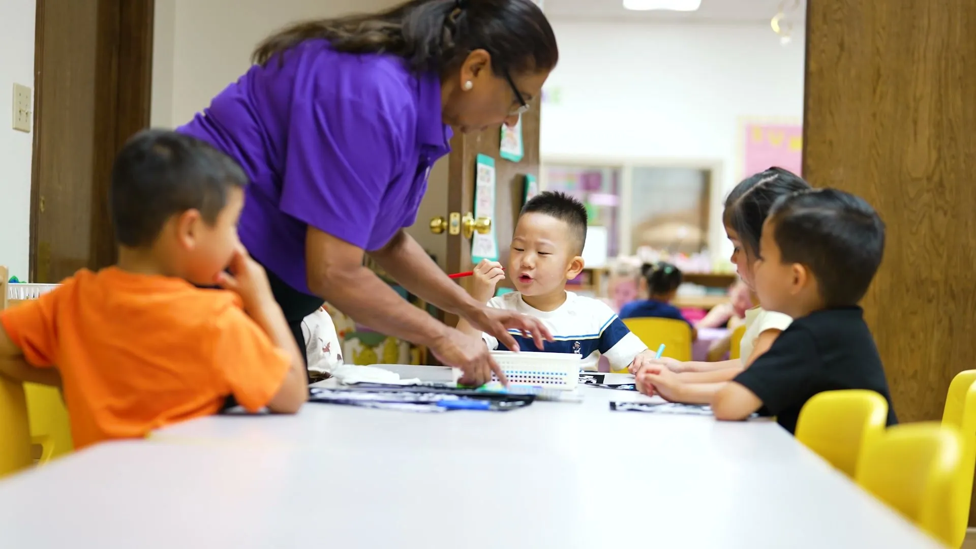 A teacher in a purple school shirt sets out materials for boys at a classroom table.