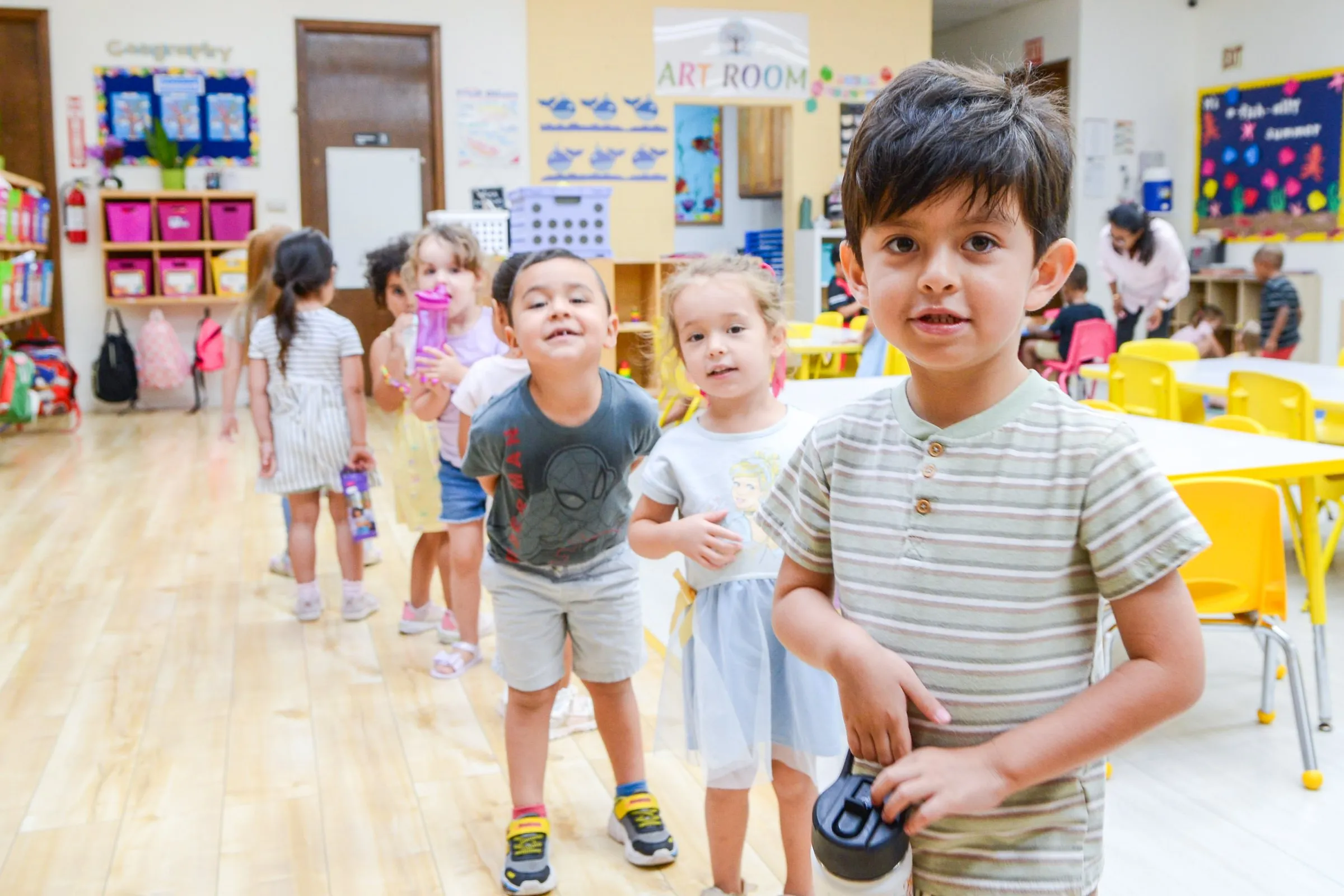 A smiling boy and his classmates walk through a bright classroom.